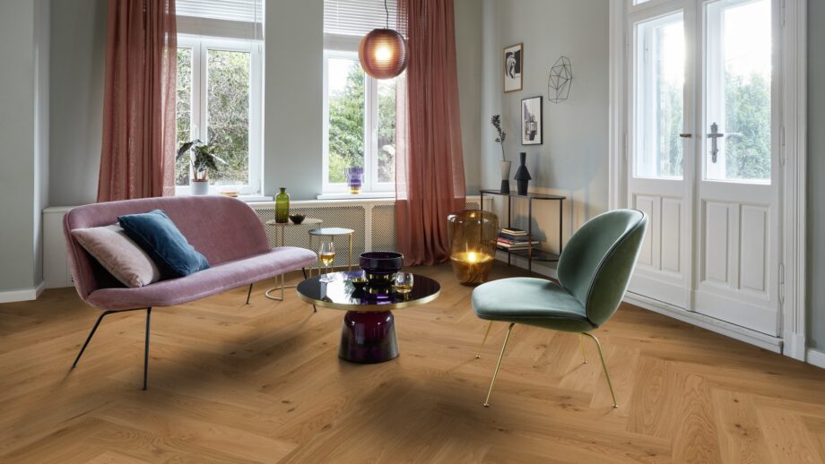 Helles, stilvolles Wohnzimmer mit Fischgrätparkett aus Eiche Mix (140 mm, geölt). Die Einrichtung kombiniert zartrosa Sofa, salbeigrünen Designstuhl und einen modernen Couchtisch mit goldenen Akzenten. Große weiße Flügeltüren und Fenster bringen viel Tageslicht in den Raum. Warme Vorhänge, moderne Kunstobjekte und eine elegante Ambienteleuchte schaffen eine harmonische und einladende Atmosphäre. Fischgrätparkett Eiche Mix 140 mm geölt im hellen Wohnzimmer mit pastellfarbenen Möbeln.
