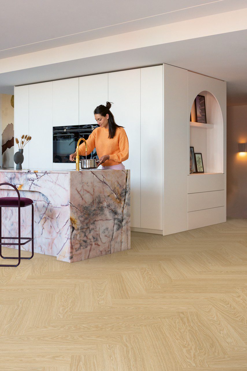 Fischgrat Vinylboden Quick Step Ciro Pure Oak Blush, verlegt auf dem Küchenboden, mit weißen Küchenmöbeln, einer Kücheninsel aus rosafarbenem Marmor mit einer goldenen Armatur, aus der eine Frau Wasser in einen verchromten Kochtopf laufen lässt.