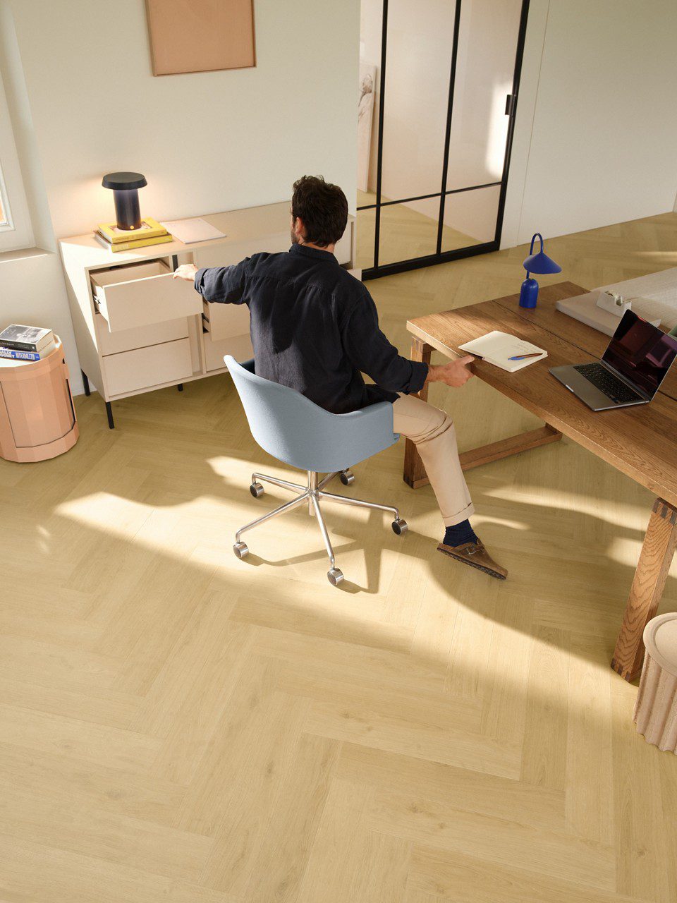 Herringbone laminate floor Quick-Step Impressive Design Nutmeg Oak installed on office floor. Guy sitting at blue chair working at office desk, laptop.