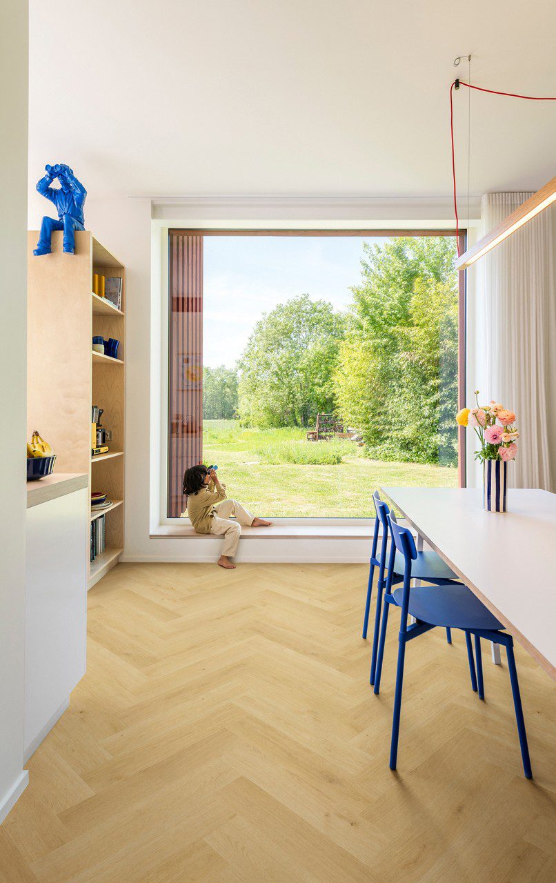 Laminate floor Quick-Step Impressive Design Nutmeg Oak installed on kitchen floor. White table with a vase on it where are flowers, blue chairs. Child with binocular looking outside of a window.