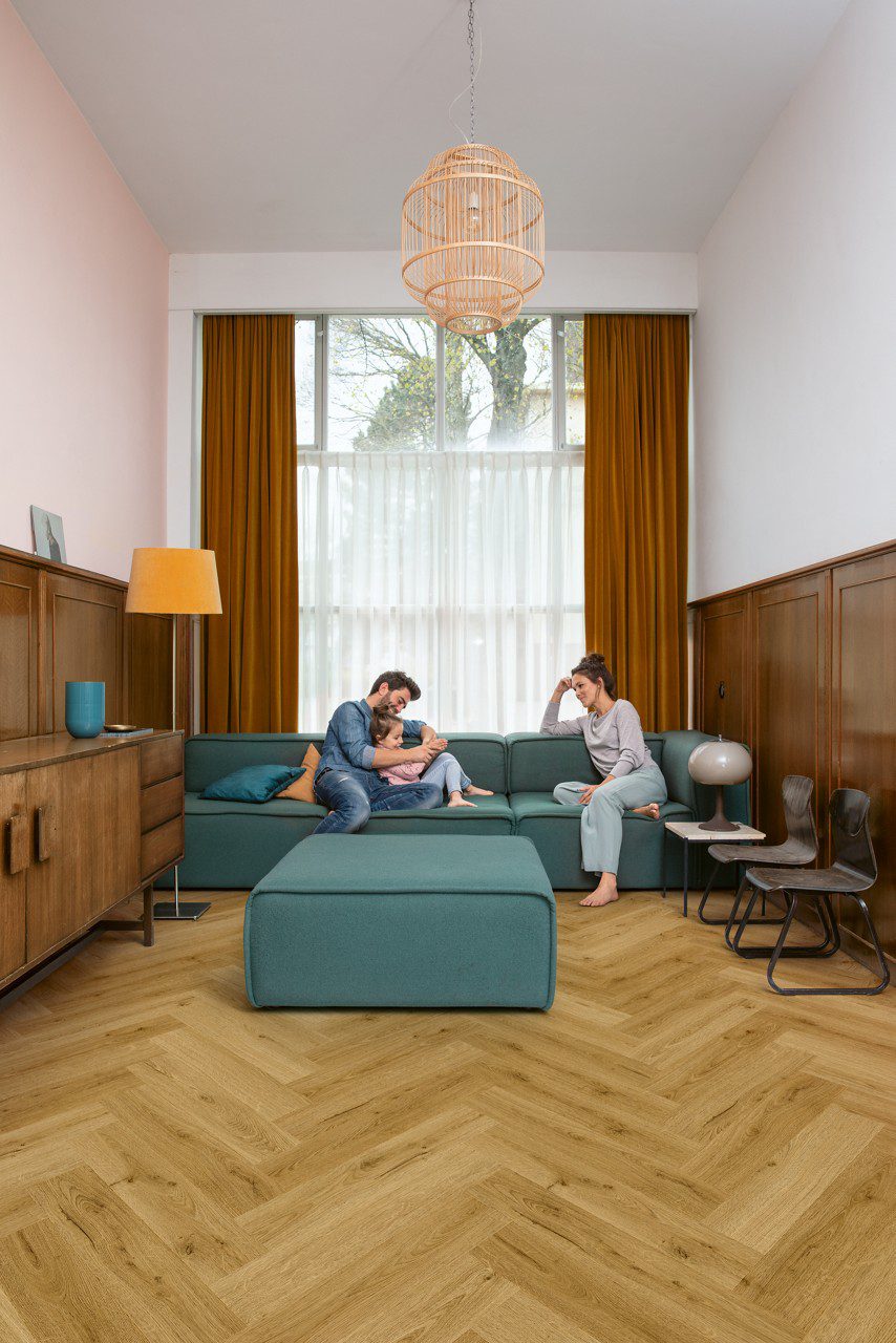 Vinyl Fischgrat Quick Step Ciro Botanic Smoked Oak, verlegt auf dem Wohnzimmerboden, ein Mann, eine Frau und ihre Tochter kuscheln auf einem blauen Sofa.