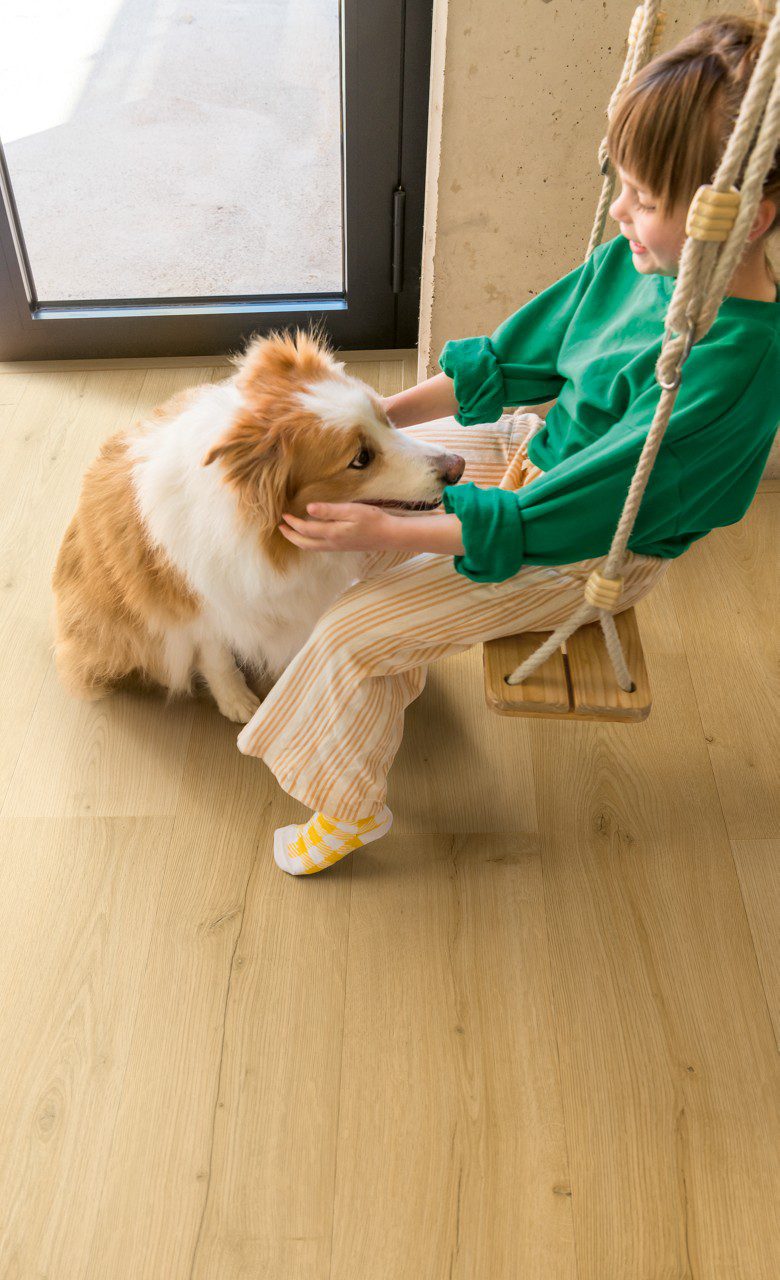 Klick Vinylboden Quick Step Bloom Brushed Oak Honey im Flur verlegt, ein Mädchen sitzt auf einer Schaukel und streichelt einen Hund.