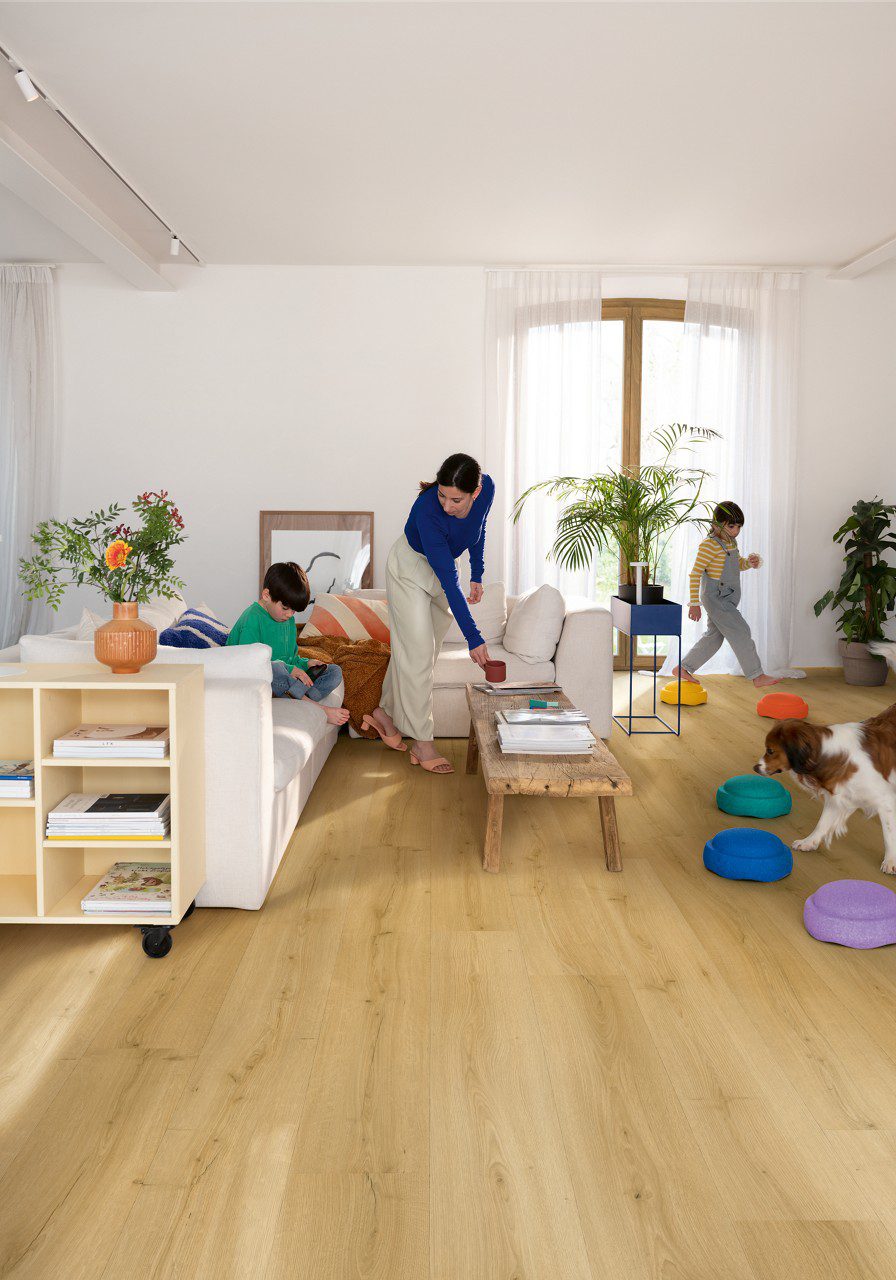 LVT parkett Quick-Step Bloom brushed oak honey paigaldatud elutoa põrandale. Ema koos kahe lapsega, koer, diican, lilled, riiul, telekalaud.