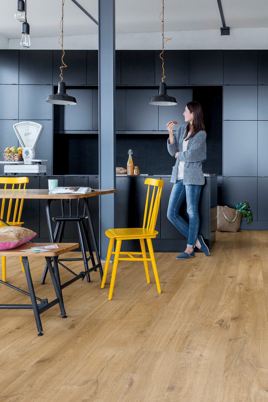 LVT parkett Quick-Step Bloom Cotton Oak Natural paigaldatud köögi põrandale. Musta fassaadiga köögimööbel, söögilaud kollaste ja mustade toolidega. Naine sööb võileiba.