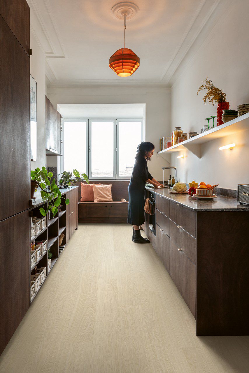 Quick Step Bloom Pure Oak Polar rigid vinyl flooring installed on a kitchen floor, a woman washing vegetables, brown kitchen cabinets.