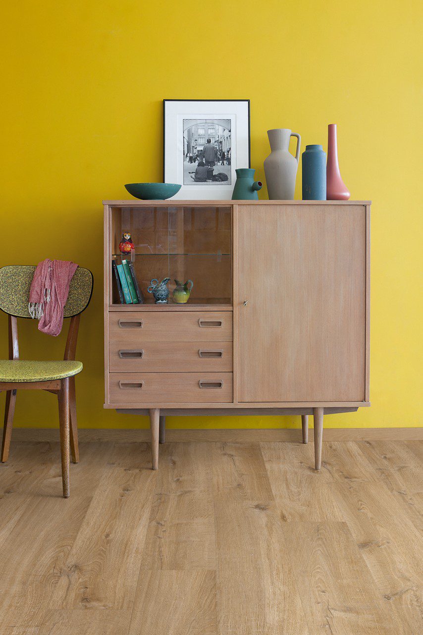 Rigid Vinylboden Quick Step Bloom Cotton Oak Natural im Wohnzimmer verlegt, gelbe Wand und klassischer Vitrinenschrank.