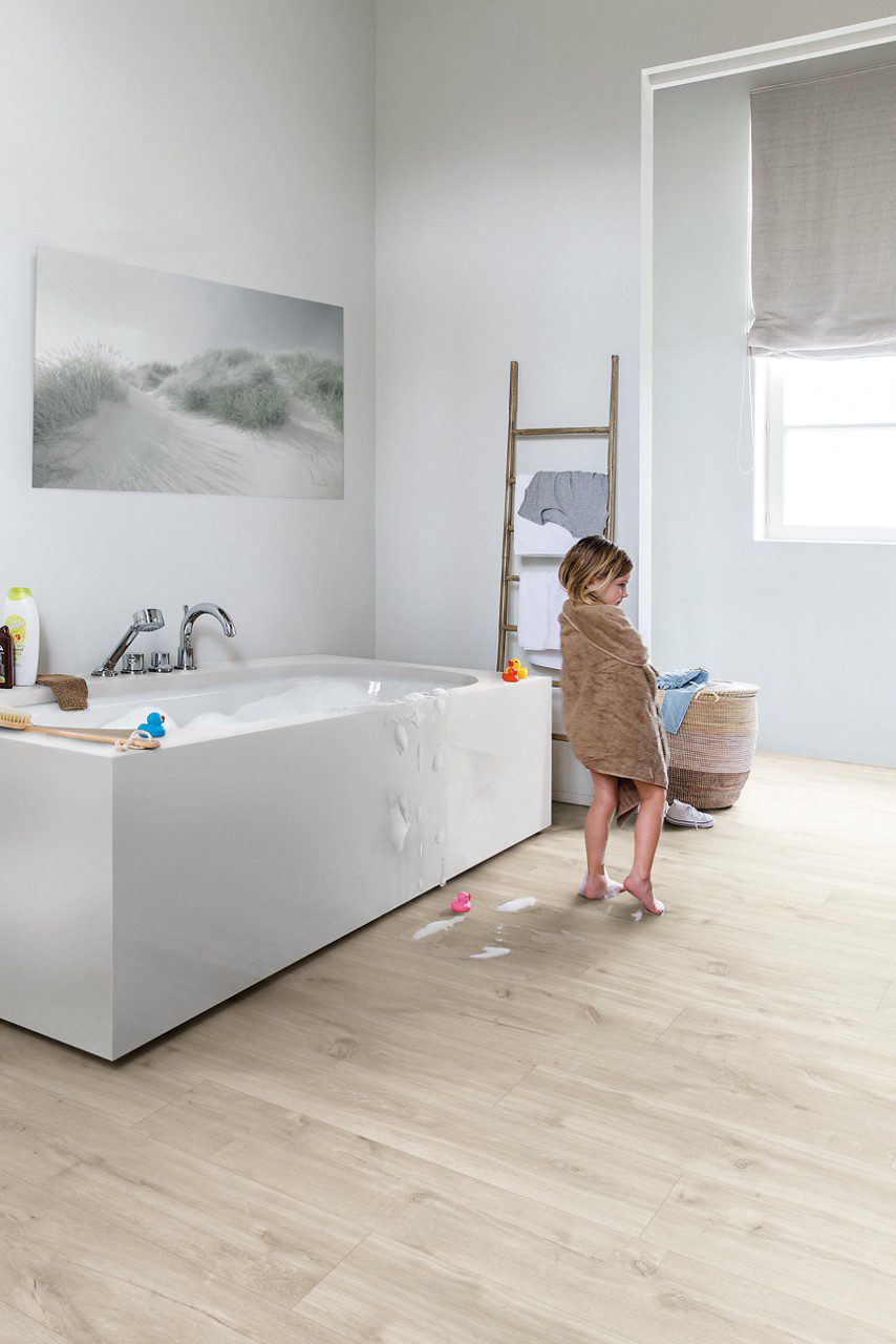 Veekindel vinüülparkett Quick-Step Blos Canyon tamm beige paigaldatud vannitoa põrandale. Valge kandiline vann on täis vannivahtu ja väike tüdruk on vannist välja tulnud ning on käterätis. Põrand on koos vannivahuse veega. Kuivaturedel millel on särk kuivamas ja seinal on pilt liivasest rannast koos kasvavate heintega.