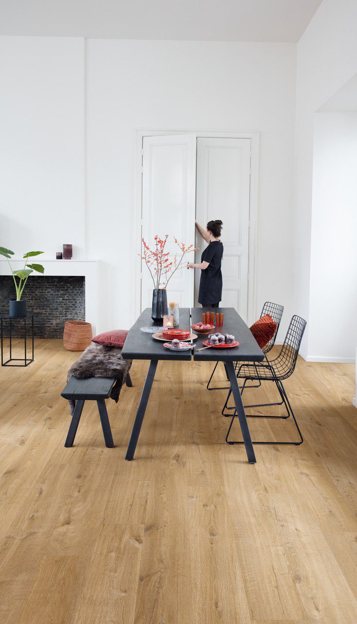 Vinüülparkett Quick-Step Bloom Cotton Oak Natural paigaldatud söögitoa põrandale. Söögilaud koos toolidega, kamina, naine võtab kapist midagi.