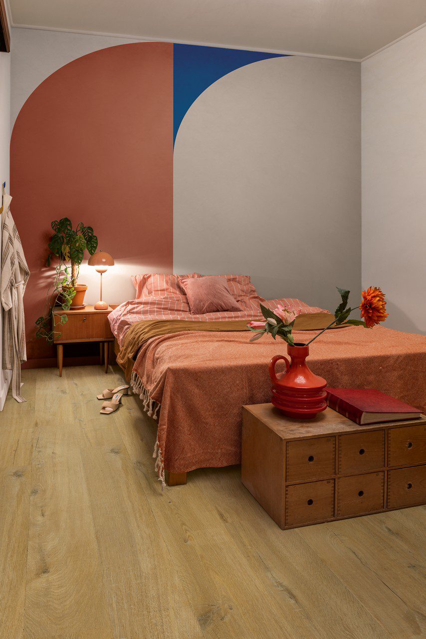 Vinüülprkett Quick-Step Bloom Cotton Oak Deep Natural paigaldatud magamistoa põrandale. Voodi, öökapp, lamp ja lill öökapil, kummut millel raamat ja vaas lilledega.