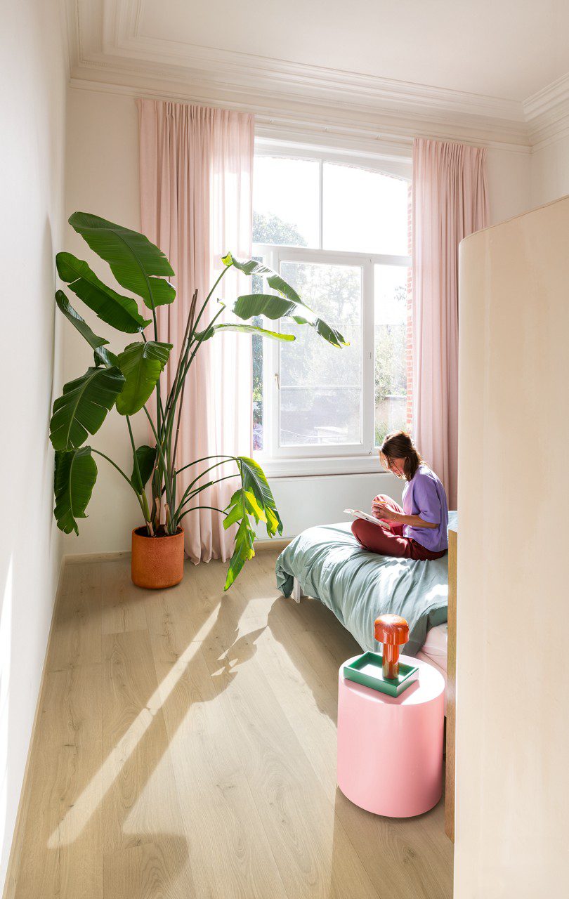 Quick Step Bloom Brushed Oak Beige vinyl floor installed in a bedroom, with a palm plant, pink curtains, and a woman reading a book on the bed.