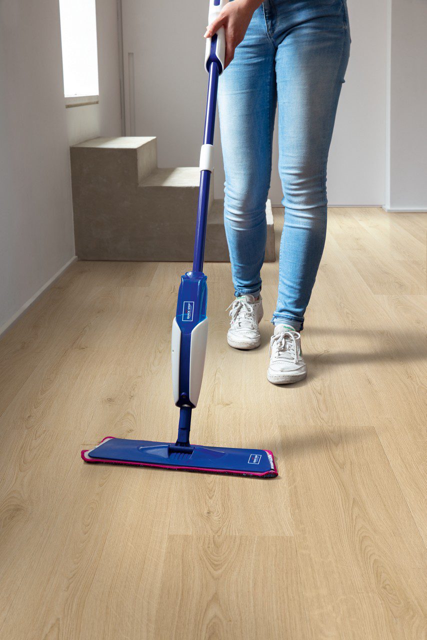 Vinylboden Quick Step Bloom Botanic Beige im Flur verlegt, eine Frau reinigt den Boden mit einem Wischmopp.