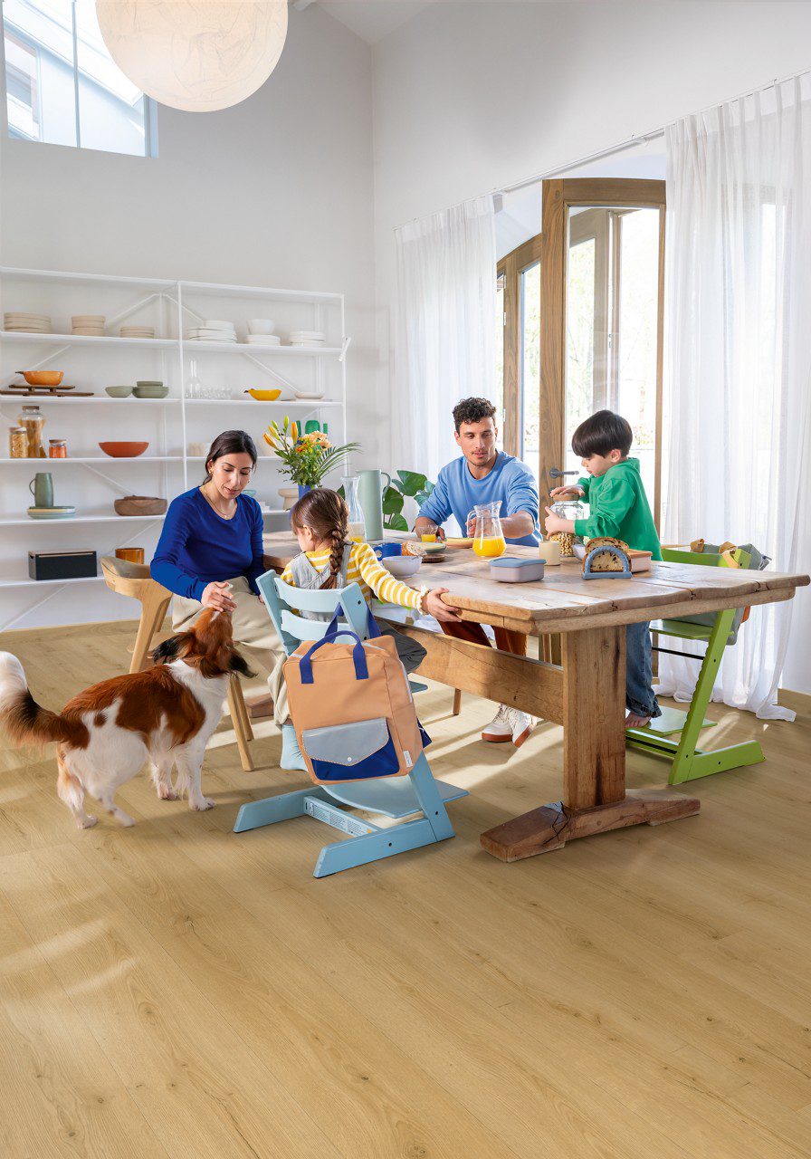 Vinyylilankku tarjous Quick Step Bloom Brushed Oak Honey asennettuna ruokailutilan lattialle, mies ja nainen syövät aamiaista lasten kanssa ruokapöydän ääressä, koira pöydän vieressä.
