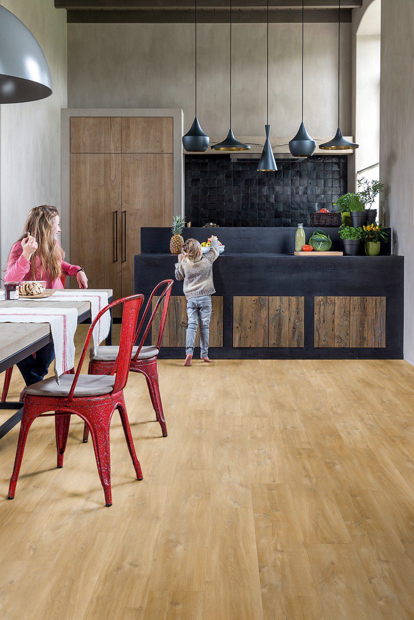 Vinyylilattia Quick Step Blos Canyon Tammi Natural asennettuna ruokailutilan lattialle, luonnollinen tammen sävy ja lankkulattia tammi, äiti istuu ruokapöydän ääressä ja seuraa tytärtä, joka ottaa banaanin hedelmävadilta keittiön kaapista.