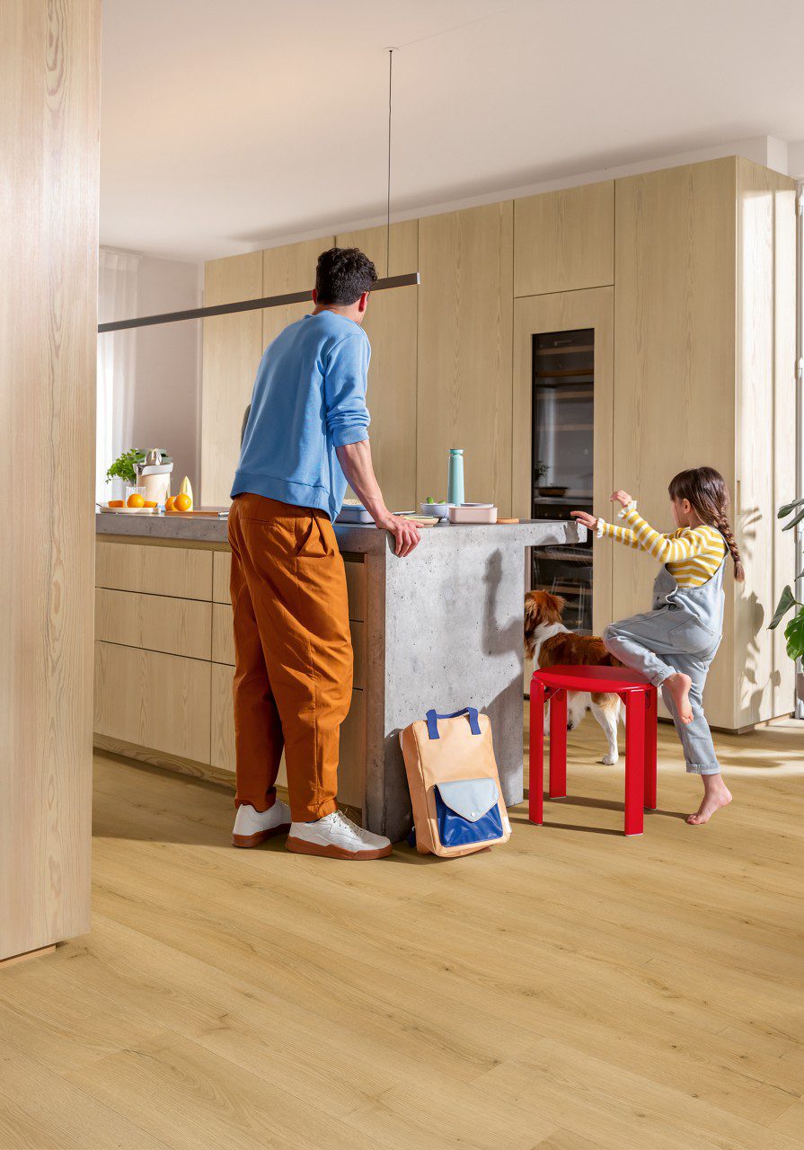 Виниловый ламинат Quick Step Bloom Brushed Oak Honey уложен на полу на кухне, отец и дочь у кухонного острова, рядом собака.