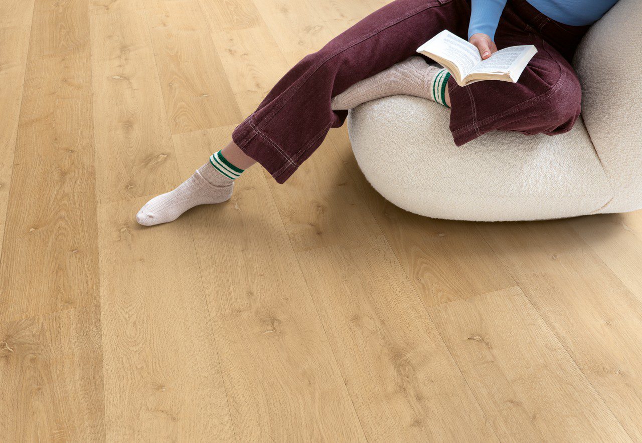 Виниловый ламинат Quick Step Bloom Elegant Oak Natural уложен на полу в гостиной, женщина сидит в удобном кресле и читает книгу.