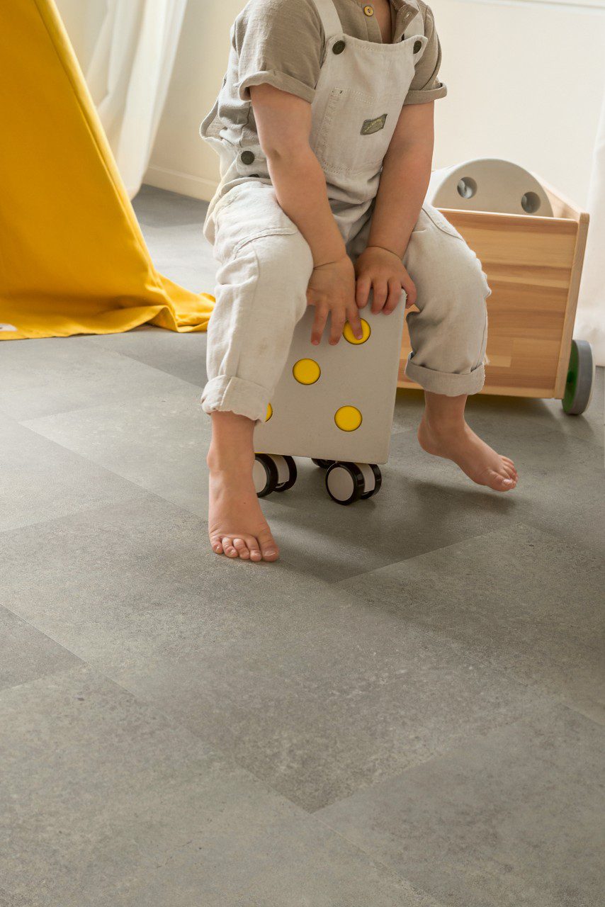 LVT parkett Quick-Step Oro Concrete rock paigaldatud lastetoa põrandale. Laps istub ratastega tumbal.