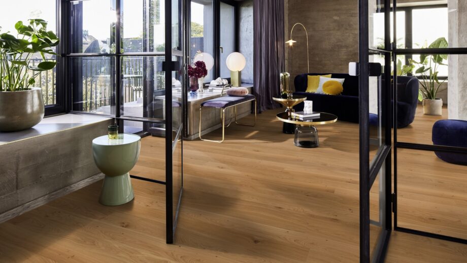 naturaalparkett-bauwerk-casapark-budget-cut-tamm-35-harjatud-olivahatatud Naturaalparkett Bauwerk Casapark Budget Cut 35 harjatud õlivahatatud naturaalse tamme tooniga ja kergelt rustikaalse sorteeringuga paigaldatud modernse elutoa põrandale. Lilla diivan, tumba ja pink. Kaks kuldset diivanilauda klaasist tasapinnaga.