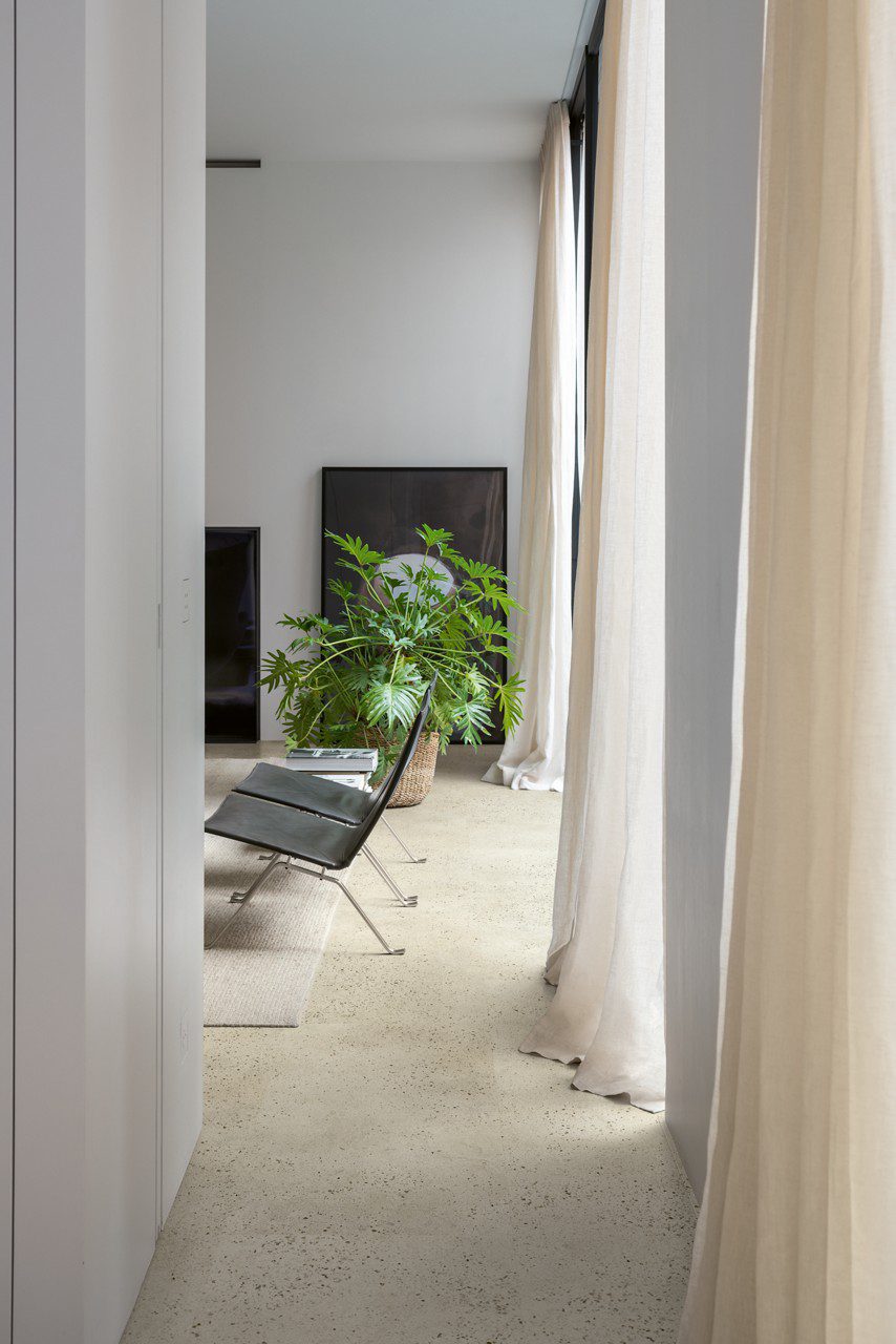 Vinylboden in Betonoptik Quick-Step Illume Pebble Concrete, verlegt auf dem Wohnzimmerboden – schwarze Lederstühle, luftige Vorhänge und eine Zimmerpflanze.