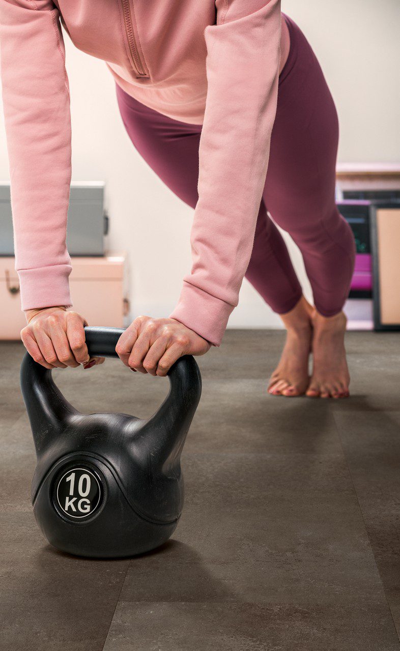 Frau macht Übungen mit einer 10-kg-Kettlebell in einem eingerichteten Home-Gym auf dem neu verlegten Vinylboden in Steinoptik Quick-Step Oro Oxidized Rock.