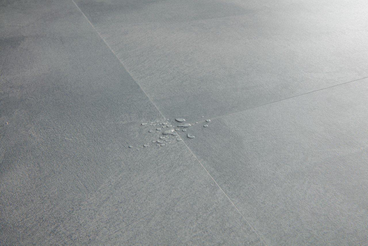 Vinyylilaatta Quick-Step Illume Soft Sky asennettuna kodin lattialle – vaalea, raikas harmaansininen sävy ja hillitty kivimäinen/betonimainen design hienovaraisella tekstuurilla; vinüylipinnan vesipisarat korostavat lattian vedenkestävyyttä.