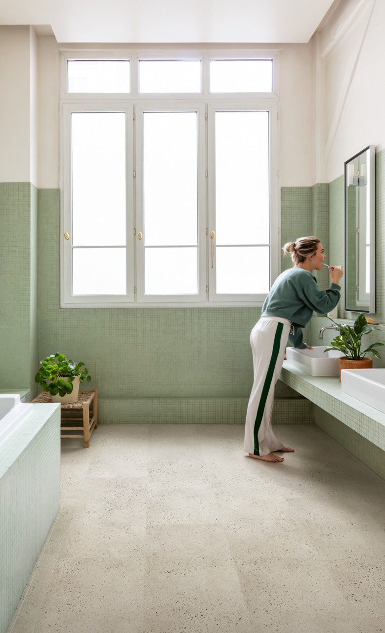 Кварц-винил Quick-Step Illume Oyster Concrete уложен на полу ванной комнаты — мебель с двумя накладными раковинами; женщина опирается рукой на раковину и чистит зубы, глядя на своё отражение в настенном зеркале.