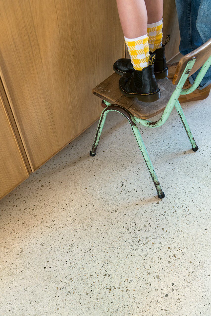 Кварц-винил Quick-Step Illume Pebble Concrete уложен на полу кухни — ребёнок стоит на старом стиле стуле, рядом стоит отец; светлый бетонный оттенок и спокойный бетонный дизайн.
