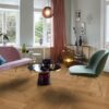 Helles, stilvolles Wohnzimmer mit Fischgrätparkett aus Eiche Mix (140 mm, geölt). Die Einrichtung kombiniert zartrosa Sofa, salbeigrünen Designstuhl und einen modernen Couchtisch mit goldenen Akzenten. Große weiße Flügeltüren und Fenster bringen viel Tageslicht in den Raum. Warme Vorhänge, moderne Kunstobjekte und eine elegante Ambienteleuchte schaffen eine harmonische und einladende Atmosphäre. Fischgrätparkett Eiche Mix 140 mm geölt im hellen Wohnzimmer mit pastellfarbenen Möbeln.
