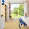 laminate-floor-quick-step-impressive-design-nutmeg-oak Laminate floor Quick-Step Impressive Design Nutmeg Oak installed on kitchen floor. White table with a vase on it where are flowers, blue chairs. Child with binocular looking outside of a window.