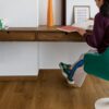 LVT flooring Quick-Step Bloom Botanic Caramel Oak installed on a living room floor. Woman is sitting on a stool.