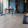 Vinyylilankku Quick Step Bloom Cotton Oak Cozy Grey asennettuna ruokailutilan lattialle, pehmeä lämminharmaa tammen sävy ja luonnollinen lajittelu, lankkudesign, siniset seinäkaapit, ruokapöytä ja eri väriset tuolit.