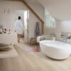 Vaalea vinyylilankku Quick Step Bloom Sea Breeze Oak Beige asennettuna kylpyhuoneen lattialle, erillinen valkoinen kylpyamme ja beige kylpyhuonekalusteet, nainen kävelee märin jaloin kylpytakissa lattialla.
