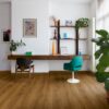 Виниловый ламинат Quick Step Bloom Botanic Caramel Oak уложен на полу в домашнем офисе, письменный стол, светло-голубое офисное кресло, пальма и цветы.